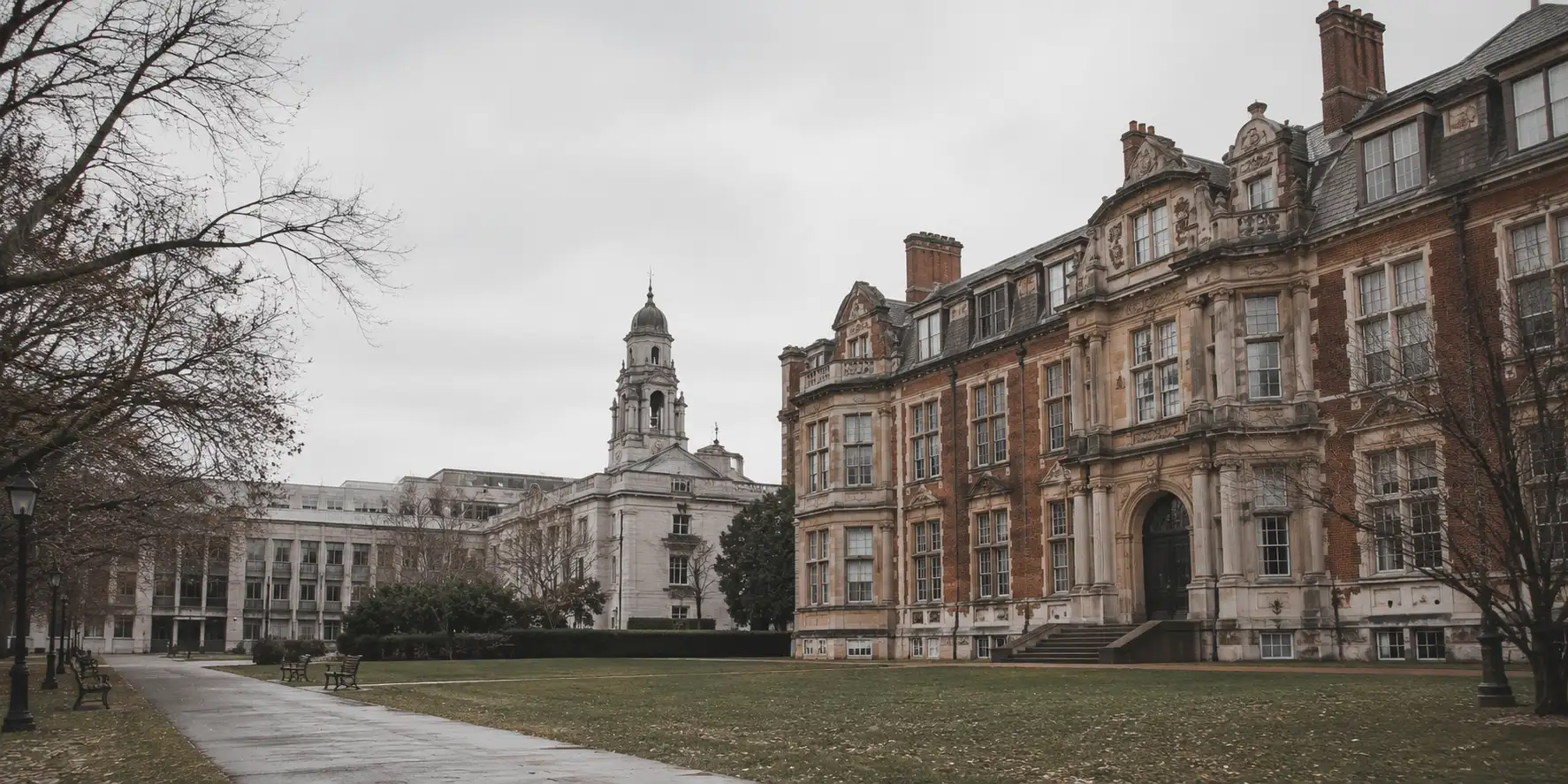 British university architecture in pale stone and red brick under overcast daylight, framed by mature trees on a quiet campus